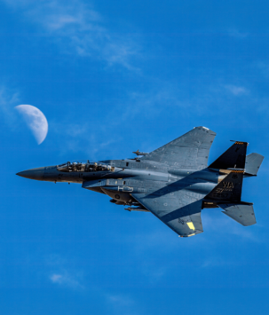 Military plane flying against a clear blue sky and half moon in the background