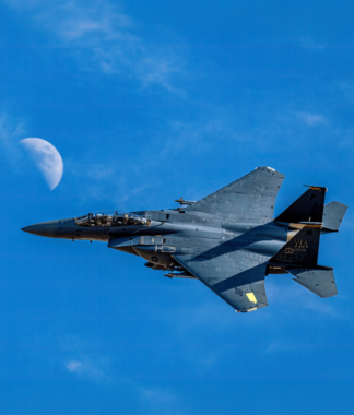 Military plane flying against a clear blue sky and half moon in the background